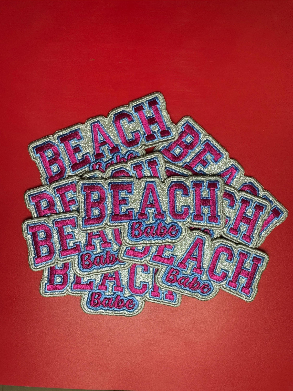 Multiple embroidered “BEACH Babe” patches with pink and blue lettering stacked together on a bright red background, showcasing bulk quantity and detailed stitching.