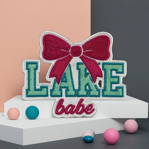 Embroidered patch with 'LAKE babe' text and a red bow on a stand with colorful balls.